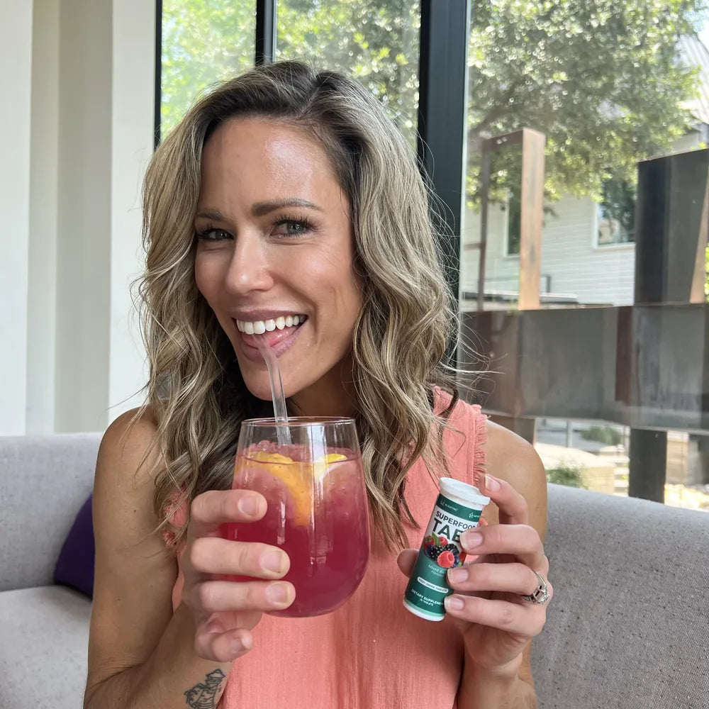 A smiling woman drinking a glass of Superfood Tabs, holding a tablet tube